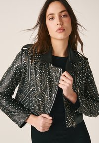 Jeune femme portant une veste en cuir noire avec des œillets argentés et des poches zippées, tenant le col de la veste sur un haut noir.