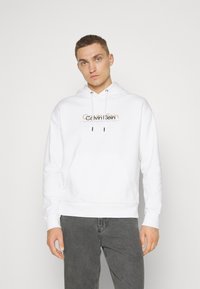 Felpa bianca con cappuccio realizzata in tessuto morbido, con tasca frontale, lacci e logo Calvin Klein in oro e nero sul petto.