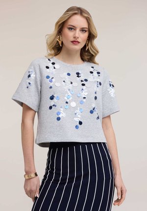 Blonde Frau trägt ein graues kurzärmeliges Oberteil mit runden Pailletten und einen schwarzen Rock mit weißen vertikalen Streifen, ergänzt durch goldene Ohrringe und ein Armband.