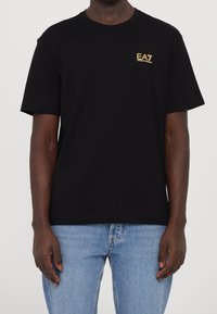 Camiseta de algodón negra con cuello redondo, mangas cortas y un pequeño logo bordado dorado en el pecho. Llevada con jeans de mezclilla de lavado claro.