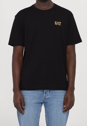 Sort bomulds-T-shirt med rund hals, korte ærmer og et lille guld broderet logo på brystet. Bæres med lysvasket denim jeans.