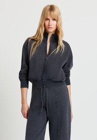 Blonde Frau trägt einen dunkel gewaschenen schwarzen Sweatshirt mit Reißverschluss und passende Jogginghosen mit Kordelzug, steht vor einem hellgrauen Hintergrund.