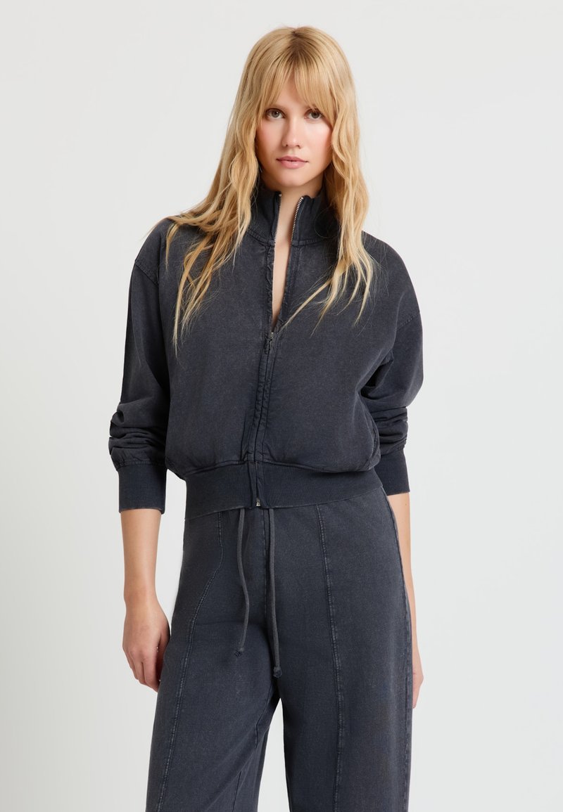 Blonde Frau trägt einen dunkel gewaschenen schwarzen Sweatshirt mit Reißverschluss und passende Jogginghosen mit Kordelzug, steht vor einem hellgrauen Hintergrund.