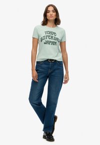 Mintgrøn t-shirt med sort grafisk tekst "TOKYO SUPERDRY JAPAN" parret med blå denimjeans. Jeansene har en lige ben-design.
