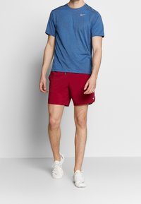 Hombre con camiseta azul de Nike, pantalones cortos deportivos rojos y zapatillas blancas caminando frente a un fondo gris liso.