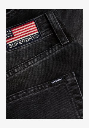 Schwarze Denim-Jeans mit einem roten, weißen und blauen gestickten Patch mit "SUPERDRY" sowie einem kleineren Etikett auf der Tasche. Strukturierter Stoff.