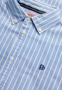 Jasnoniebieska koszula na guziki z białymi pionowymi paskami, zapinanym kołnierzykiem i małym granatowym haftowanym logo na piersi.