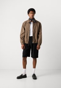 Hnedá ľahká bomber bunda, biele tielko, čierne šortky, čierne slip-on topánky a čierny šál, stojí na jednoduchom pozadí.
