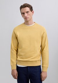 Mann mit kurzem, lockigem Haar, der einen schlichten gelben Pullover über einem weißen Hemd und dunklen blauen Jeans trägt, steht vor einem schlichten, hellen Hintergrund.