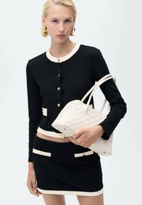 Cardigan nero a maglia con rifiniture crema, bottoni dorati, abbinato a una gonna nera con tasche crema. Tiene una borsa intrecciata bianca.