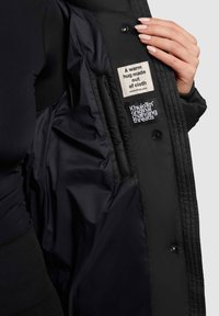 Giacca nera imbottita con una texture liscia. L'interno presenta due etichette: un'etichetta in tessuto beige e un logo ricamato nero.