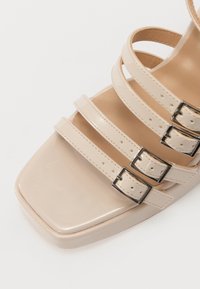 Sandale en cuir verni beige avec un bout carré, dotée de quatre fines lanières et de trois accents de boucle en argent. Texture lisse et design épuré.