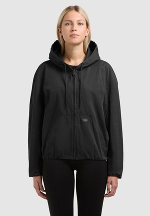 Femme aux cheveux blonds lisses portant une veste noire à capuche zippée avec des poches frontales et un pantalon noir, debout devant un fond uni.