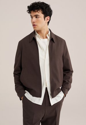 Junger Mann mit lockigem Haar, der eine dunkelbraune Jacke mit Reißverschluss über einem weißen, gestreiften Hemd mit Knöpfen trägt und die Hände in den Taschen hat.