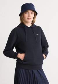 Sudadera de color azul marino hecha de una tela suave, que cuenta con un bolsillo frontal, capucha con cordón y un pequeño logo verde. Combinada con una falda de tela azul marino con pliegues.
