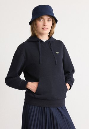 Marineblå hettegenser laget av mykt stoff, med en frontlomme, snor i hetta og en liten grønn logo. Kombinert med et plisserte marineblå skjørt.
