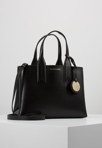 Svart läderhandväska med dubbla handtag, strukturerad yta, spetsiga detaljer, guldfärgad logotyp och avtagbar metallcharm. Inkluderar avtagbar rem.
