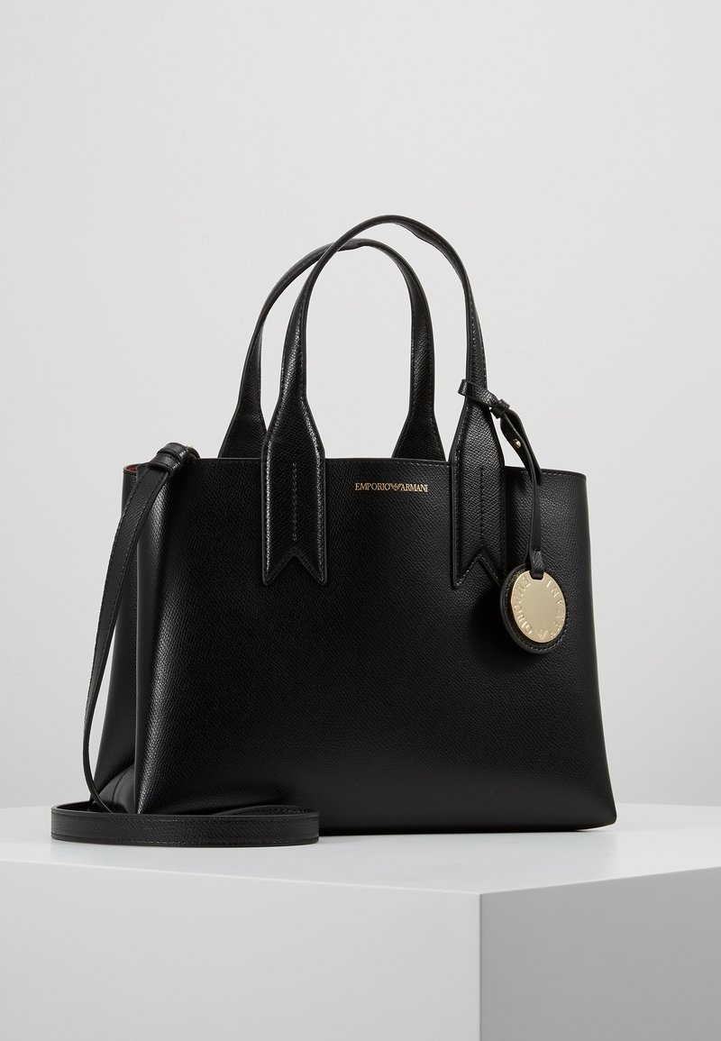 Svart läderhandväska med dubbla handtag, strukturerad yta, spetsiga detaljer, guldfärgad logotyp och avtagbar metallcharm. Inkluderar avtagbar rem.