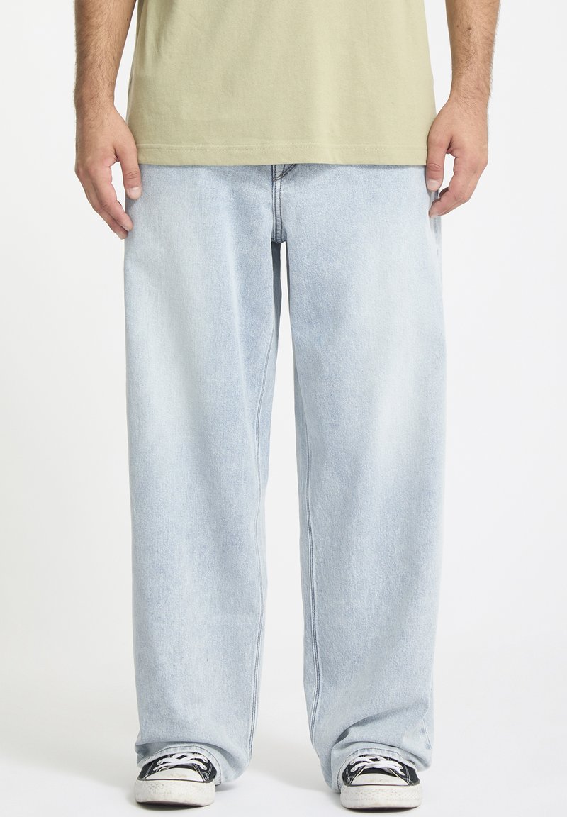 Hellblaue Jeans aus Denim mit lockerer Passform, geraden Beinen und einem ausgewaschenen Finish. Getragen mit schwarzen Sneakers mit weißen Akzenten.