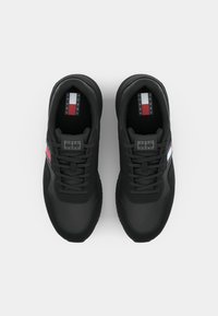 Schwarze Sportschuhe aus strukturiertem Stoff, mit Schnürdesign und blauen Nähten. Die Seite weist rot-, weiß- und blau gestreifte Akzente auf.