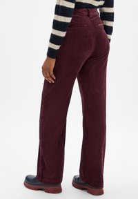Pantalons en velours côtelé bordeaux avec des côtes verticales, taille haute et poches arrière. Le modèle porte des chaussures noires à semelles épaisses.