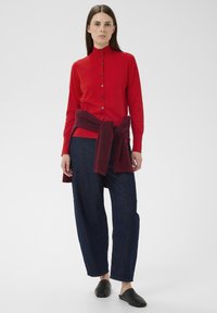 Pulover roșu cu nasturi și mâneci lungi, asortat cu pantaloni lejeri din denim închis și pantofi slip-on negri, pulover visiniu legat la talie.