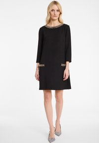 Femme blonde portant une robe noire longueur genou avec un col en perles et des détails de poches à l'avant, associée à des escarpins argentés à bout pointu.