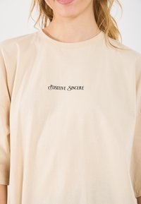 Beige katoenen t-shirt met korte mouwen, met de tekst "Positive Sincere" in contrasterende zwarte letters aan de voorkant.