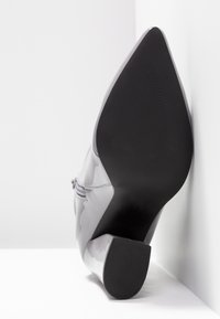 Bottine blanche brillante à talon, inclinée vers le haut, montrant la semelle pointue noire et le talon bloc sur fond blanc.