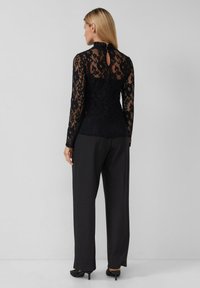 Haut en dentelle noire à manches longues, col haut et détail en forme de clé à l'arrière, associé à un pantalon large noir et des chaussures à talons.