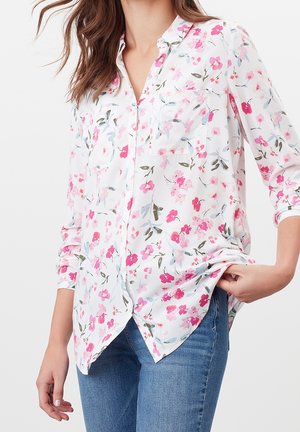 Kvinde iført hvid bluse med lange ærmer og pink blomsterprint samt blå jeans, holder i blusens kant med den ene hånd.