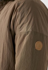 Giacca leggera marrone con motivo a griglia, realizzata in un tessuto testurizzato e con un logo circolare in pelle color tan sulla manica sinistra.