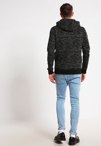 Svart kamuflasje hettegensere med romslig hette, ribbede mansjetter og kant, kombinert med lyseblå jeans og svarte sko.