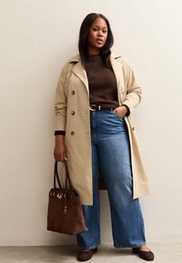 Un abrigo trench beige con botones de doble botonadura sobre un suéter marrón, combinado con jeans de pierna ancha azules y un bolso tote de ante marrón.