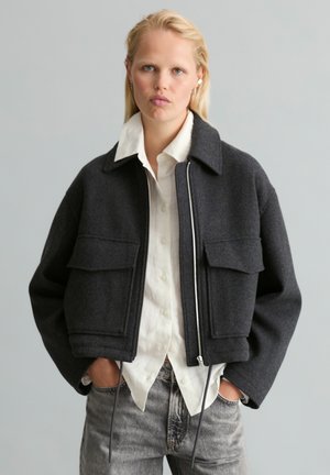 Femme blonde portant une veste courte gris foncé sur une chemise blanche boutonnée et un jean gris, debout les mains dans les poches devant un fond uni.
