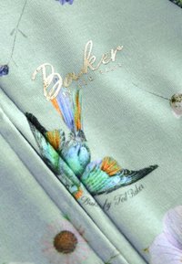 Svijetlozeleni materijal s cvjetnim uzorkom i ilustracijom šarene leptirice. Zlati logo "Baker by Ted Baker" integriran u dizajn.