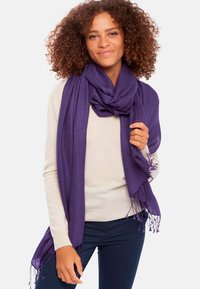 Écharpe violette en matériau doux, avec des bords frangés et un design drapé. Le modèle la porte sur un pull léger.