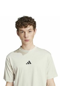 Camiseta de algodón color beige claro con mangas cortas, que presenta un pequeño logo negro de Adidas en el pecho y un escote redondo.