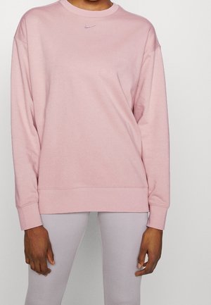 Persoon draagt een lichtroze Nike-trui en lichtgrijze legging, staand tegen een effen witte achtergrond.