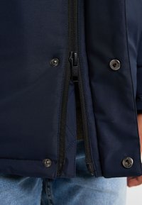 Giacca blu navy con zip frontale, bottoni a scatto e foderatura interna testurizzata. Presenta doppie zip e tessuto liscio e resistente all'acqua.