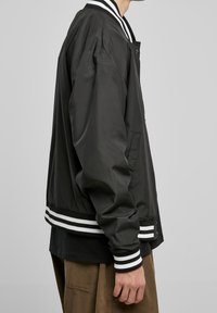 Czarna kurtka typu bomber o gładkiej fakturze, z białymi paskami na kołnierzu i mankietach oraz bocznymi kieszeniami, pokazana na osobie z boku.