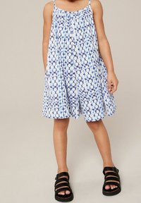 Dziecko w bezrękawowej, sięgającej do kolan białej sukience z niebieskim wzorem tie-dye oraz czarnych sandałach z klamrą, stoi na jednolitym tle.