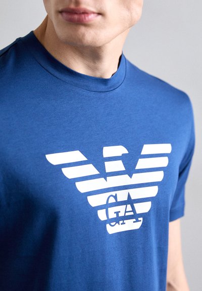 Sinine puuvillane T-särk ümmarguse kaelusega, millel on valgete triipudega logo kujundus, kus tõstetuna on näha "GA" logo all.