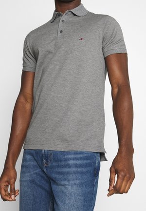 Mand iført en grå kortærmet poloshirt med et lille rødt, hvidt og blåt logo på brystet og blå jeans.