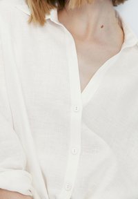 Witte linnen blouse met een V-hals, knoopsluiting en opgerolde mouwen. De stof heeft een lichte, gestructureerde uitstraling.