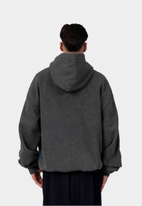 Sweat à capuche gris, coupe ample, avec des poignets côtelés. Fabriqué dans un tissu texturé et possède une petite étiquette bleue sur le côté gauche.