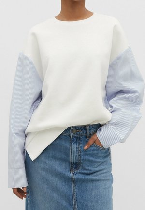Personne portant un haut blanc à manches longues rayées bleu clair et une jupe en denim à taille haute, de couleur délavée moyenne, avec un bouton et une fermeture éclair à l'avant.