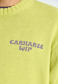 Pull en tricot vert clair avec un col côtelé. Présente le texte "CARHARTT WIP" en violet, mettant en valeur un tissu texturé.