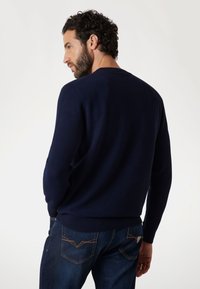 Maglione lavorato a maglia blu navy con maniche lunghe; polsini e orlo a coste; indossato con jeans blu scuro con cuciture a contrasto sulle tasche.