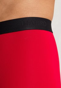 Leggings rossi con una texture liscia e una vita elastica nera con logo in rilievo. Si concentra su un design aderente e aerodinamico.